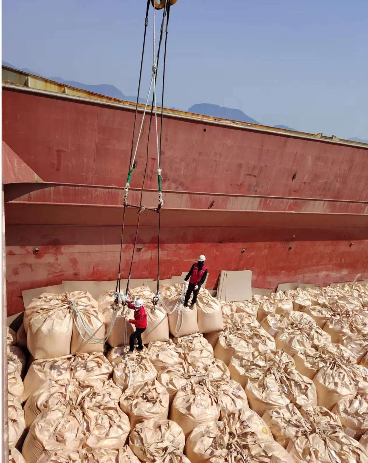 Workers rigging slings on FIBC jumbo bags beside a vessel